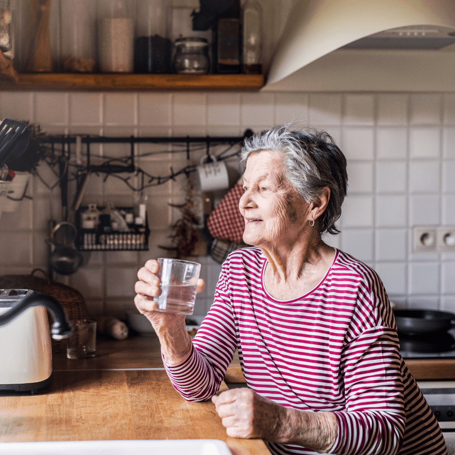elderly woman looking out a window in a clean kitchen drinking water 